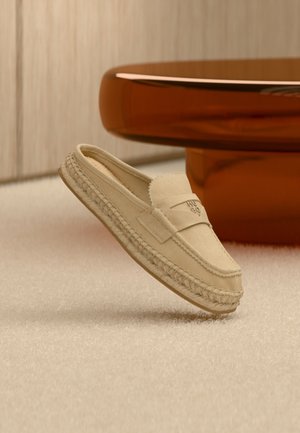 Mocassin beige en daim à enfiler avec semelle tressée et logo embossé, posé sur un tapis clair près d'un pied de table rond en bois.