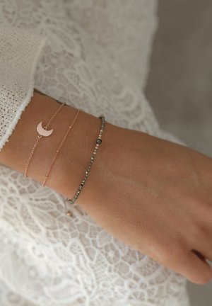 Tre fine armbånd på et håndled: et charm i rosa guld formet som en halvmåne, et slankt kæde i rosa guld, og et perlearmbånd med grå og sølvtoner.