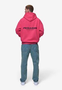 Rozā kapuces džemperis ar melnu "PEGA DOR" uzrakstu, apvienots ar gaiši ziliem džinsiem un baltām kedām. Audums ir maigs un nedaudz teksturēts.