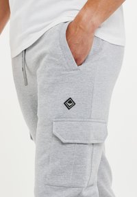 Pantalon de jogging gris en tissu doux, avec une poche cargo sur le côté, une taille élastique et un petit écusson noir sur la cuisse.