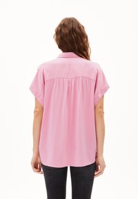 Pink, short-sleeved blouse with a soft, shiny texture. Features a button-down collar and pleating at the back. Worn with dark jeans.