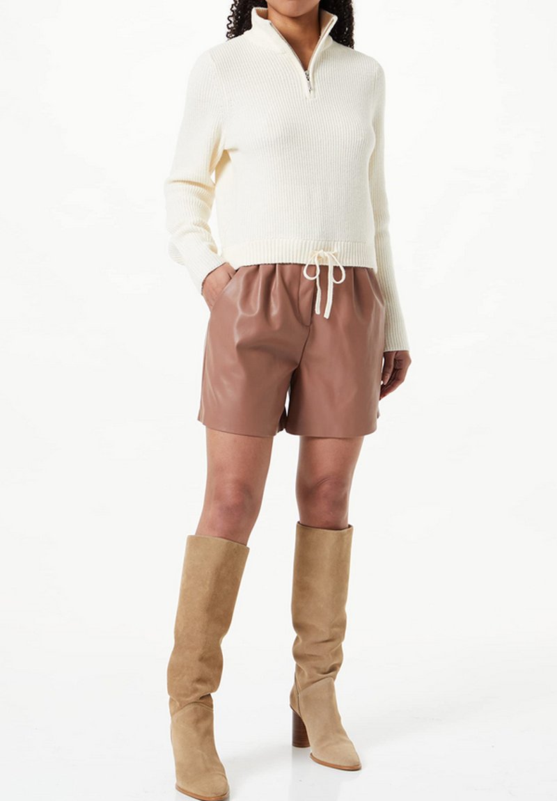 Sweater de malha creme com gola com fecho, combinado com calções de pele castanhos e botas de camurça bege até ao joelho com um salto grosso.
