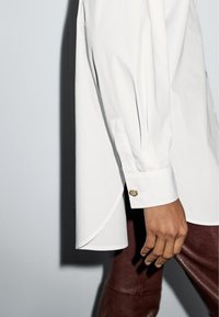 Bílá košile s dlouhými rukávy, zakulaceným lemem a zlatými knoflíky na manžetách. Kombinována s tmavě hnědými kalhotami se strukturovaným vzorem.