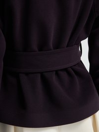 Blazer viola scuro con cintura con dettagli plissettati, texture in tessuto morbido e vestibilità su misura, indossato sopra abbigliamento beige.