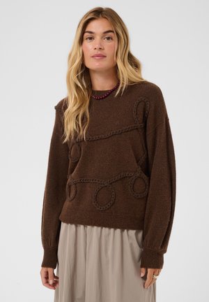 Femme blonde portant un pull marron avec des motifs de boucles tressées et une jupe beige sur un fond clair uni.