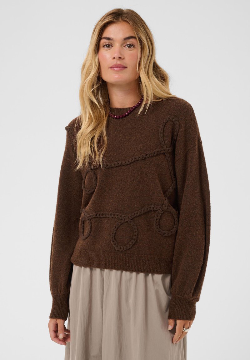 Femme blonde portant un pull marron avec des motifs de boucles tressées et une jupe beige sur un fond clair uni.
