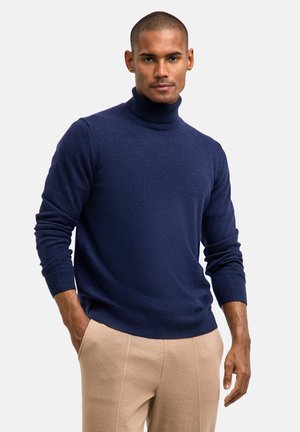 Pull col roulé bleu marine en tissu doux, doté de manches longues et de poignets côtelés. Assorti à un pantalon beige clair, le mannequin se tient avec confiance.