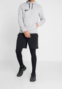 Homme portant un sweat à capuche Nike gris clair, un short Nike noir par-dessus un legging noir, des chaussettes noires et des chaussures de sport noires, debout devant un fond blanc.