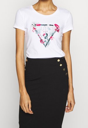 Femme portant un t-shirt blanc à manches courtes avec un logo floral Guess et une jupe noire taille haute avec des boutons dorés sur le côté.