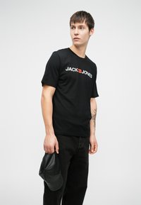 Melna kokvilnas T-kreklu ar "JACK & JONES" logo baltā un sarkanā krāsā. Modelis tur melnu ādas cepuri. Vienkāršs apaļš kakla izgriezums, īsās piedurknes, brīvs griezums.