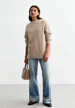 Beiger Kabelstrickpullover mit Rundhalsausschnitt, kombiniert mit hellblauen Schlagjeans und schwarzen Ballerinas. Das Model hält eine kleine beige Handtasche.