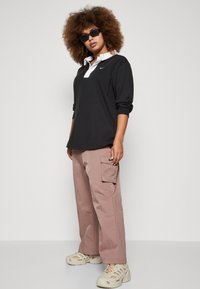 Polo noir à manches longues avec un col blanc, associé à un pantalon cargo rose clair. Le modèle porte des lunettes de soleil noires et des chaussures beiges.