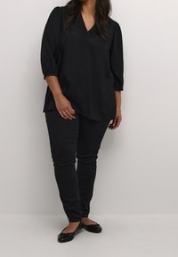 Blouse noire ample avec un col en V et des manches trois-quarts, associée à un pantalon noir slim et des chaussures plates. Texture lisse.