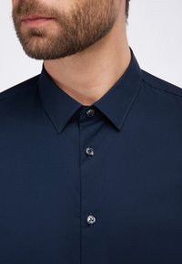 Camisa azul marino de botones con cuello clásico, fabricada en una tela suave y con cuatro botones en el frente, que muestra un diseño elegante y ajustado.