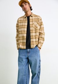 Brunternetternet flannel skjorte med en brystlomme, båret over en sort skjorte. Parret med løse blå denim jeans med sidelommer. Brun kasket.