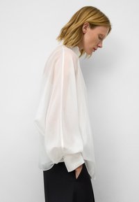 Doorzichtige witte blouse met lange, luchtige mouwen en een hoge kraag. Soepele textuur met een losse pasvorm. Gecombineerd met donkere broek.