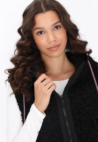Schwarze Fleece-Weste mit Reißverschluss und pinken Kordelzügen, kombiniert mit einem weißen Langarmoberteil mit strukturiertem Material.