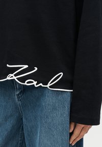 Μαύρο πουκάμισο με μακρύ μανίκι και λευκή κεντημένη υπογραφή «Karl» στο κάτω μέρος, συνδυασμένο με ανοιχτόχρωμο μπλε τζιν παντελόνι. Ορατή απαλή υφή υφάσματος.