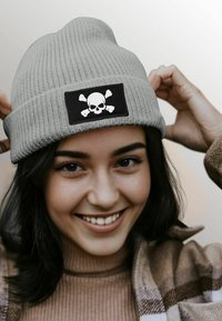 Gray ribbed knit beanie featuring a black patch with a white skull and crossbones graphic. Two hands adjusting the hat are visible.