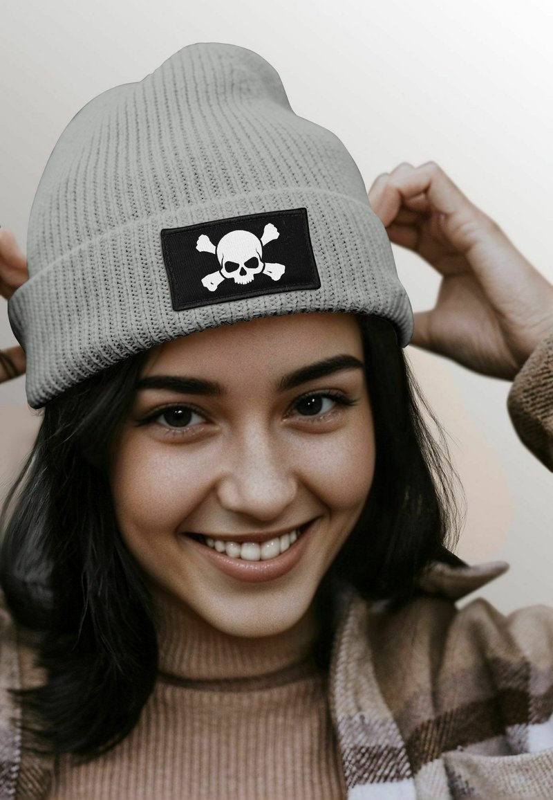 Gray ribbed knit beanie featuring a black patch with a white skull and crossbones graphic. Two hands adjusting the hat are visible.