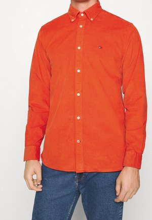 Mand iført en lyseorange kordfløjls-skjorte med knapper og krave samt blå jeans, stående mod en ensfarvet baggrund.