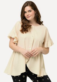Blusa corta beige chiaro con maniche a sbuffo, apertura con bottoni e orlo svasato. Indossata sopra pantaloni neri a motivi con forme geometriche.