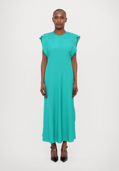 Une longue robe turquoise avec des manches courtes, fabriquée dans un tissu léger, présentant une coupe droite et une texture lisse. Des chaussures noires à talons pointus complètent le look.