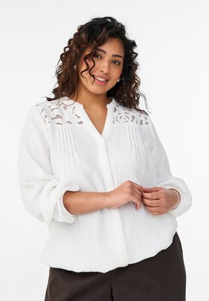 Ženska z kodrastimi lasmi, oblečena v belo bluzo z reliefno teksturo in čipkastimi detajli na ramenih, z zavihanimi rokavi in temnimi hlačami, nežno se smehlja.