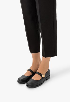 Chaussures Mary Jane noires avec boucle ronde dorée portées avec un pantalon noir court sur pieds nus sur fond blanc.