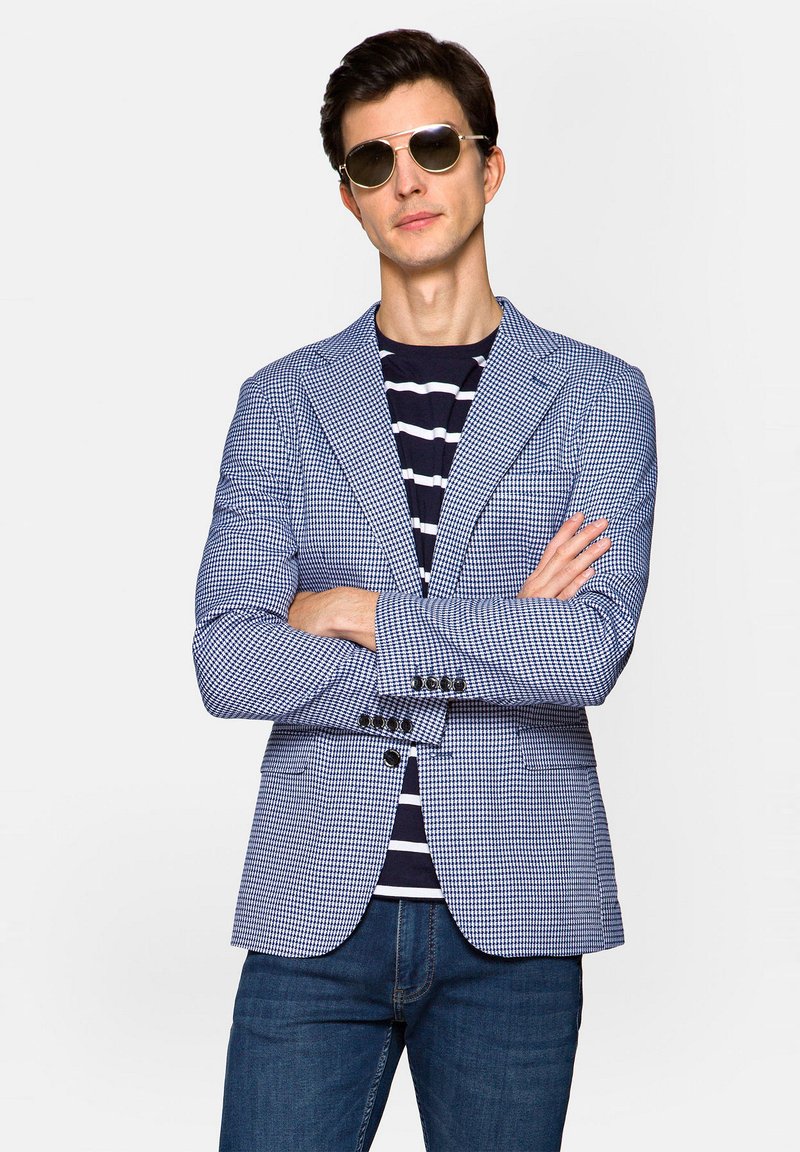 Niebiesko-biały marynarka w kratkę z dwoma guzikami, klapami o prostokątnym kształcie i dobrze dopasowanym kroju, noszona na granatowej koszuli w paski. Uzupełniona ciemnymi dżinsami.