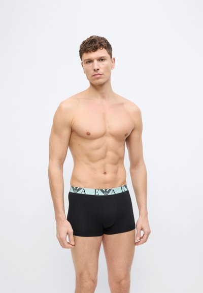 Ανδρας χωρίς μπλούζα με κοντά, σγουρά μαλλιά, φορώντας μαύρα μπόξερ με μπλε ζώνη μέσης, στέκεται μπροστά σε απλό λευκό φόντο.