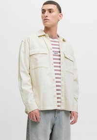 Man in een roomkleurig corduroy overshirt over een mauve en witte gestreepte t-shirt, lichtblauwe spijkerbroek, kijkend naar links tegen een effen achtergrond.