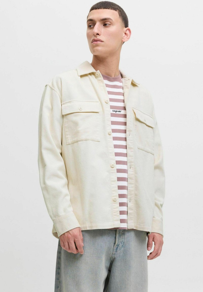 Man in een roomkleurig corduroy overshirt over een mauve en witte gestreepte t-shirt, lichtblauwe spijkerbroek, kijkend naar links tegen een effen achtergrond.