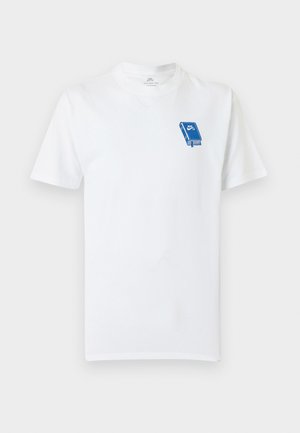 Młoda osoba ubrana w biały t-shirt z niebieskim logo, luźne czarne spodnie, białe trampki oraz jasnoniebieską czapkę z daszkiem, stoi z rękami w kieszeniach.