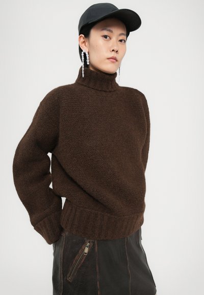 Brauner Strickpullover mit hohem Kragen und geripptem Saum, aus strukturiertem Stoff. Kombiniert mit einem schwarzen Lederrock mit Reißverschluss und einer Mütze.
