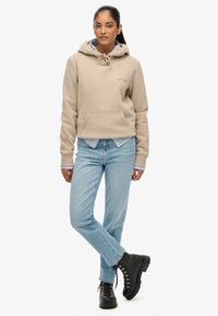 Felpa beige con tasca a marsupio, cordoncino regolabile e logo ricamato. Jeans azzurri con una sottile texture denim, abbinati a stivali neri con lacci.