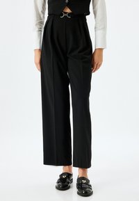 Pantalons noirs taille haute avec des plis, accompagnés d'une ceinture à boucles argentées. Assortis à une chemise blanche à manches longues et des chaussures noires.
