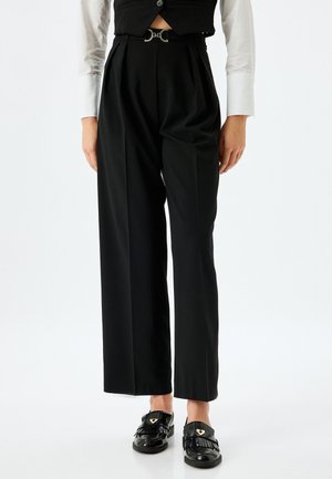 Black, high-waisted trousers with pleats, completed with a silver buckle belt. Paired with a white long-sleeve shirt and black shoes.