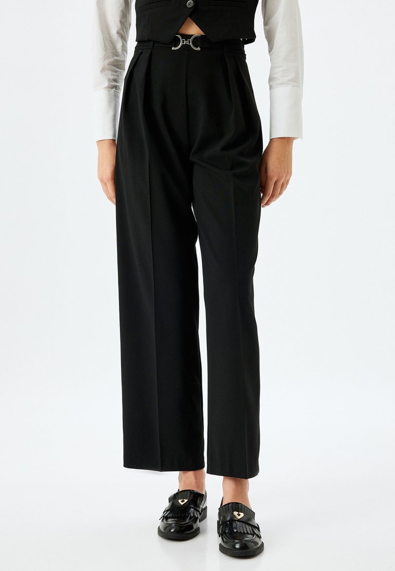 Pantalons noirs taille haute avec des plis, accompagnés d'une ceinture à boucles argentées. Assortis à une chemise blanche à manches longues et des chaussures noires.
