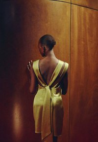 Robe en satin jaune avec un dos en V profond, présentant un détail de taille nouée et des accents noirs contrastants, sur fond en bois.