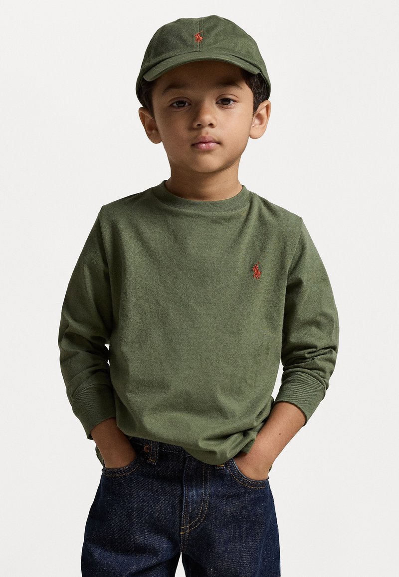 Zöld, hosszú ujjú póló kerek nyakkal és kis piros logóval, zöld kalappal párosítva, sötétkék farmernadrággal viselve.