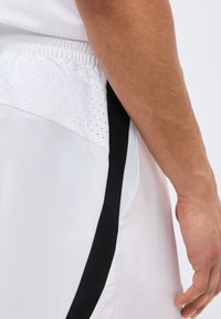 Ανδρικός βραχίονας δίπλα σε λευκά αθλητικά σορτς με μαύρη κάθετη ρίγα και διάτρητο πάνελ κοντά στη λαστιχένια ζώνη της μέσης.