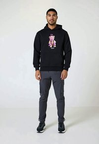 Hoodie preto com uma gráfica colorida de urso, combinado com calças cargo cinzas e sapatos desportivos pretos. Tecido suave, ajuste casual, design versátil.