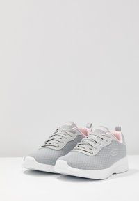Zapatillas deportivas grises con parte superior de malla texturizada, suela blanca y acentos en rosa claro en el cuello y la lengüeta. Presentan un sistema de cordones tradicional.