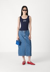 Body din navy cu text "GAP", fustă maxi din denim albastru cu buzunare laterale, pantofi roșii cu talpă joasă și o poșetă mică albastră.
