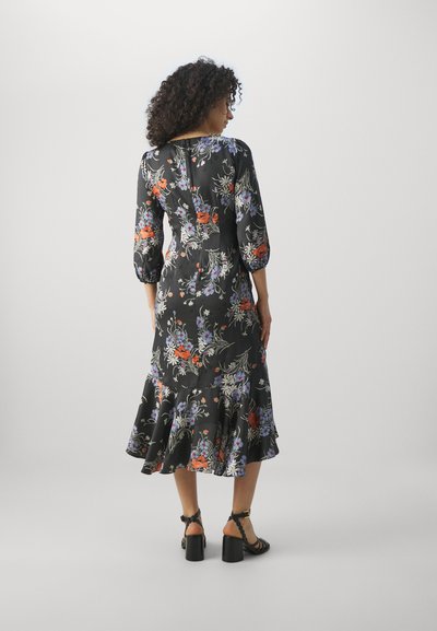 Schwarzes Blumenkleid mit Dreiviertelärmeln, rüschendem Saum, mit bunten Blumen in Rot, Blau und Weiß. Getragen mit schwarzen Absätzen.