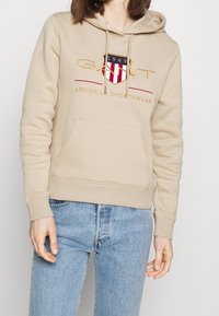 Felpa beige con tasca frontale, cappuccio con cordino e logo ricamato con dettagli rossi e blu. Tessuto morbido e polsini a costine.