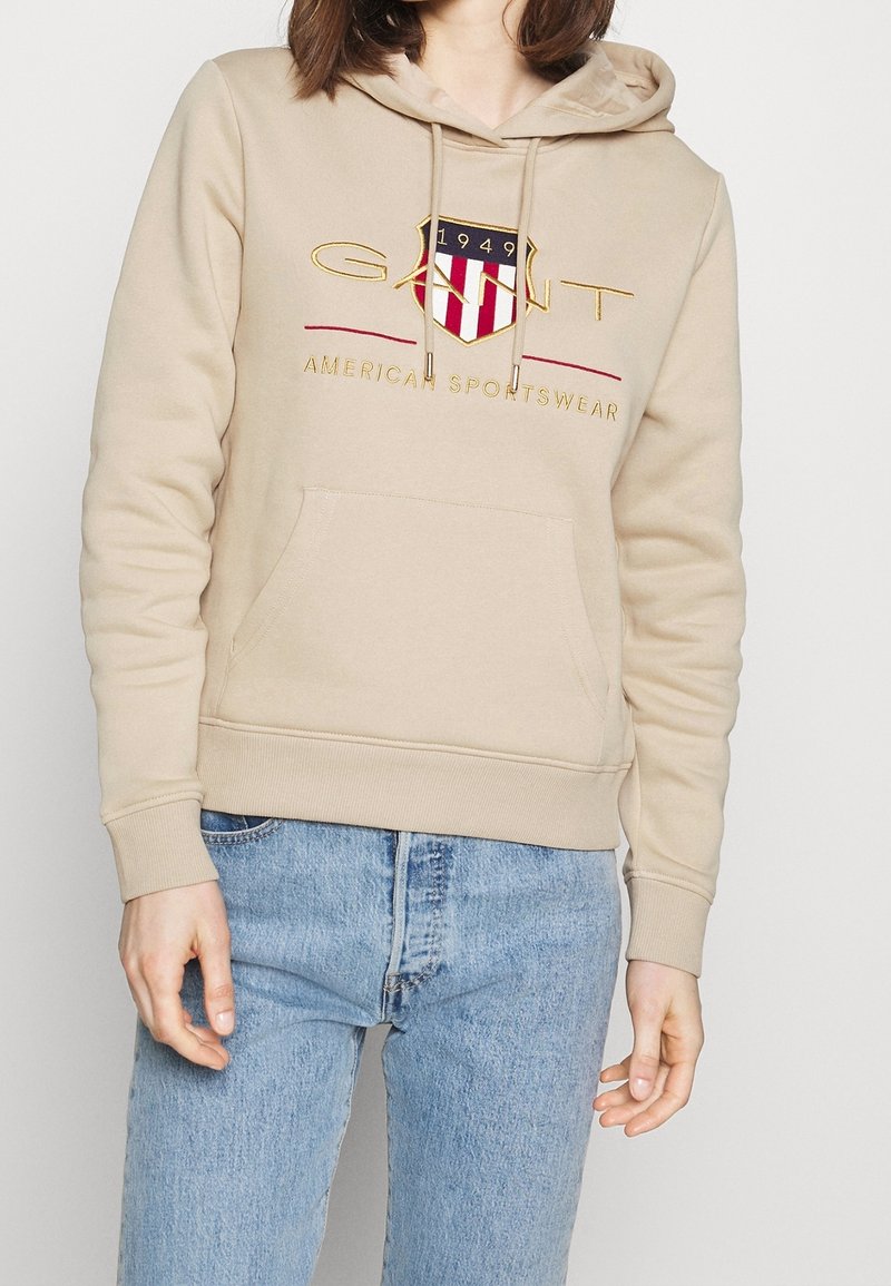 Sudadera beige con un bolsillo frontal, capucha con cordones y logo bordado que presenta acentos en rojo y azul. Tejido suave y puños acanalados.