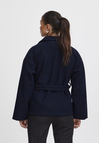 Giacca in pile blu navy con vita cinturata, maniche ampie e colletto. Texture liscia e linee pulite, adatta per look sovrapposti.
