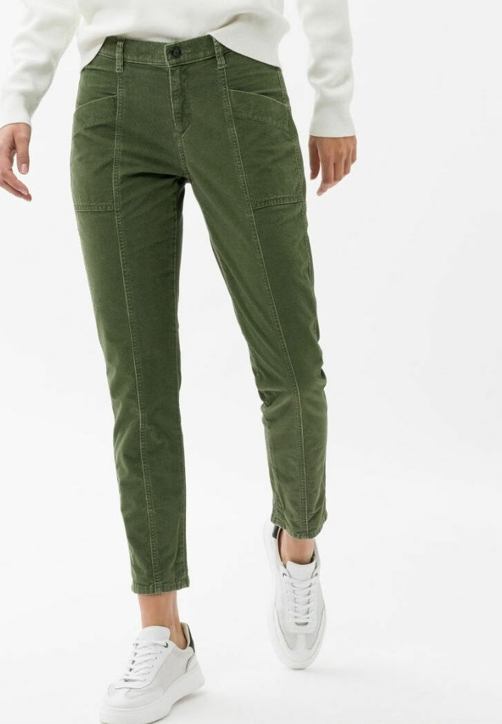 Personne portant un pantalon slim vert olive, un pull blanc et des baskets blanches, debout sur un fond blanc.